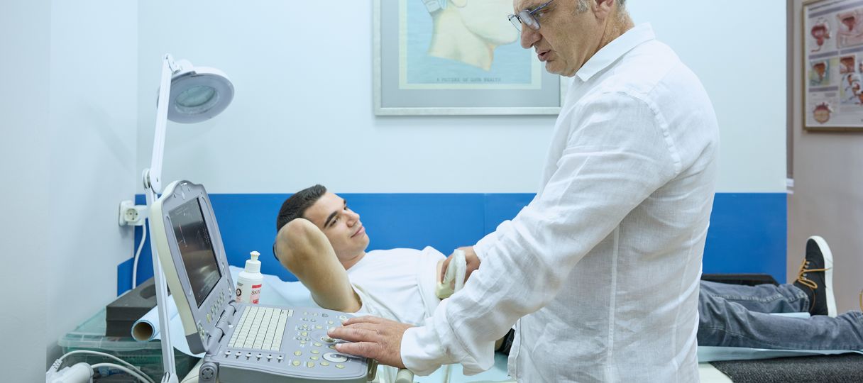 Η φωτογραφία δείχνει τον ουρολόγο Οδυσσέα Σοπηλίδη να εξετάζει τον ασθενή με τη χρήση υπερηχογραφήματος. Ο ασθενής είναι ξαπλωμένος σε ένα εξεταστικό κρεβάτι και έχει σηκώσει την μπλούζα του, αποκαλύπτοντας την κοιλιά του για την εξέταση. Ο ουρολόγος κρατάει τον υπερηχογραφικό ανιχνευτή και παρακολουθεί την οθόνη της συσκευής υπερήχων για να ελέγξει τα αποτελέσματα. Ο ασθενής φαίνεται χαλαρός και άνετος κατά τη διάρκεια της εξέτασης. Στον τοίχο υπάρχει ένα διάγραμμα του ανθρώπινου εγκεφάλου, το οποίο διακοσμεί τον χώρο.